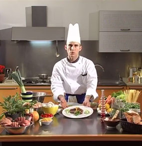 Un cuoco mostra un piatto sul tavolo di una cucina dove sono presenti molti ingredienti colorati tra cui notiamo delle parti umane come mani e orecchie