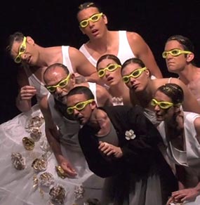 Un gruppo di attori con abiti bianchi tranne quello in primo piano con giacca nera e occhiali da piscina gialli
