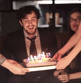 Un ragazzo sorride illuminato solo dalle candeline di una torta che gli viene avvicinata da quattro mani