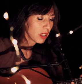 Anna Vignozzi canta un brano live con chitarra e occhi chiusi, in esterni notturni con lucine di atmosfera